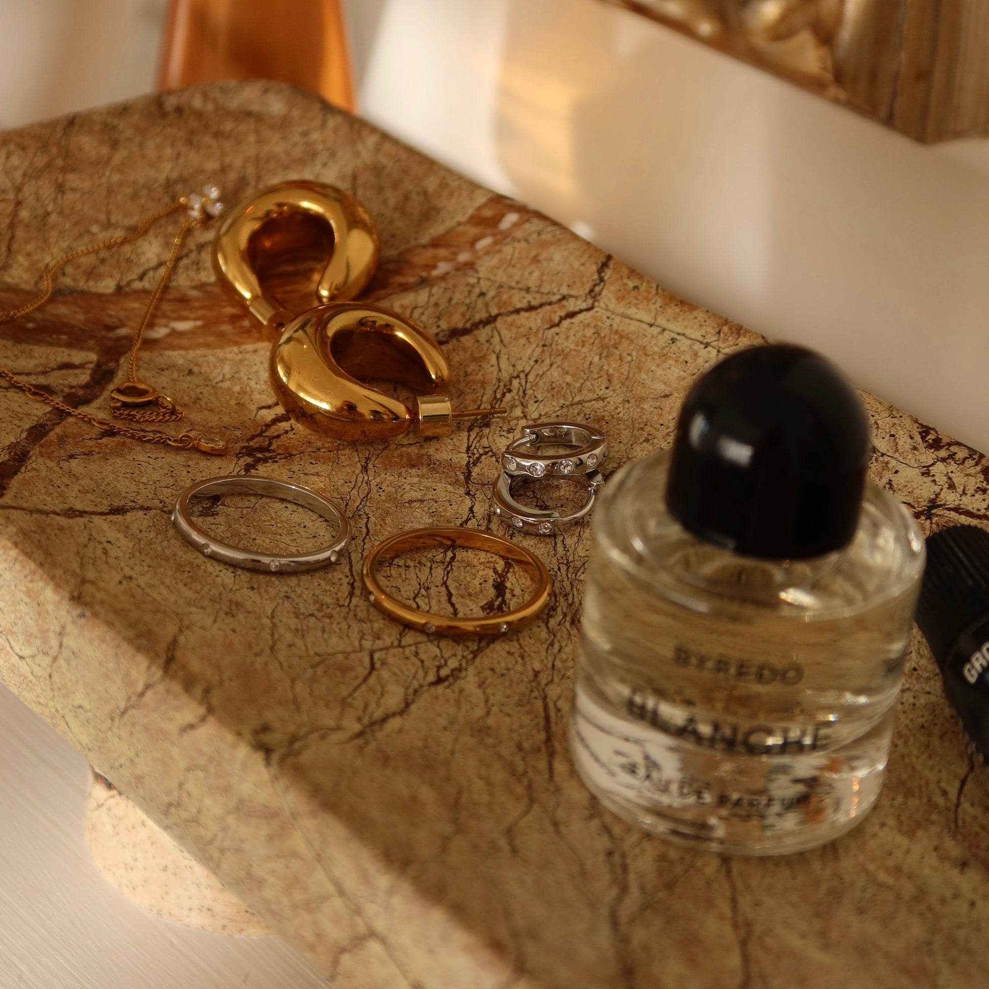 Gold earrings and rings on a marble surface with a Byredo perfume bottle.